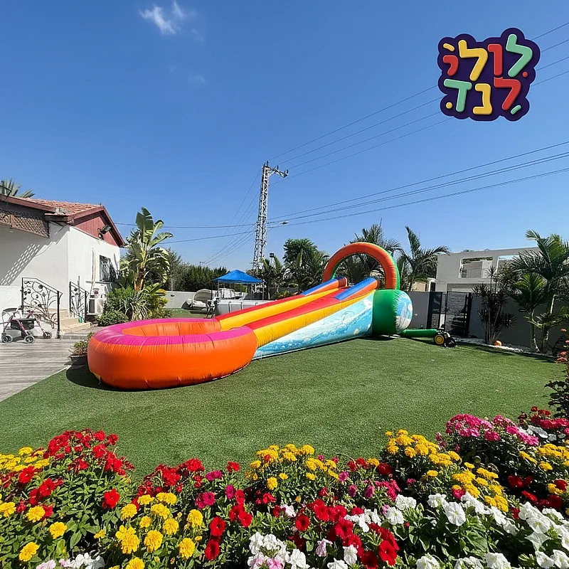 מגלשת קצף מתנפחת ארוכה בצבעי כתום, צהוב וכחול, עם בריכת נחיתה עגולה, מוצבת על דשא סינתטי בחצר של בית פרטי